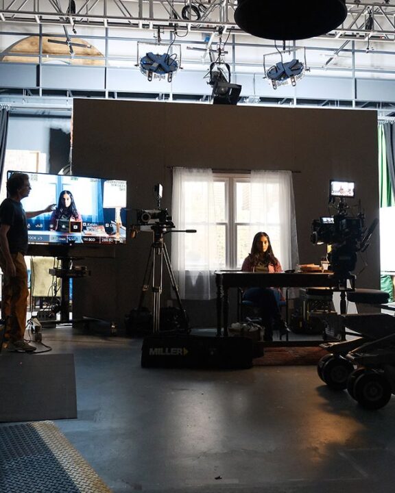 A soundstage with an actor sitting at a table. In the foreground are cameras and monitors.