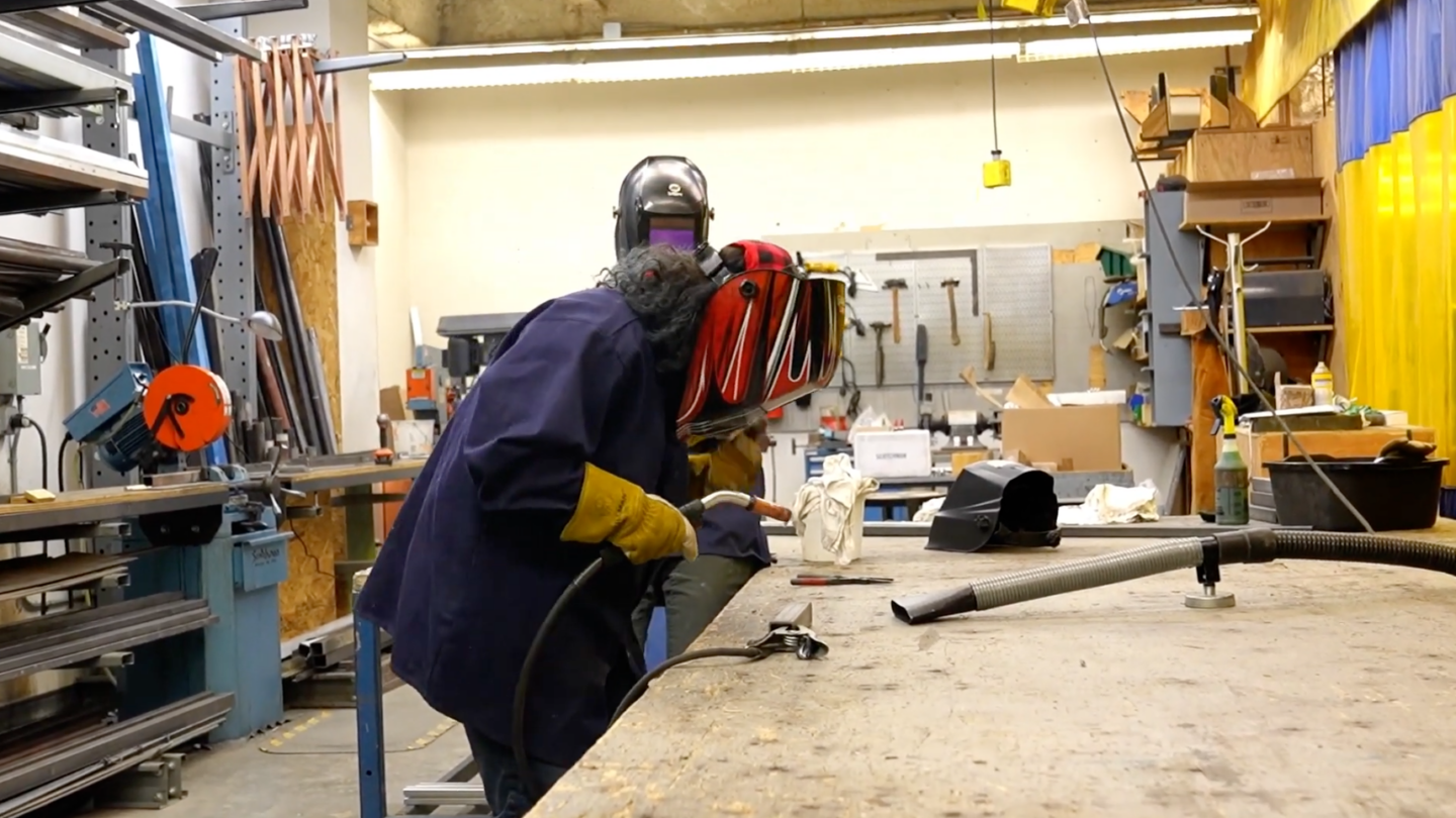 A student with a mask on leans over a work piece with a welding tool