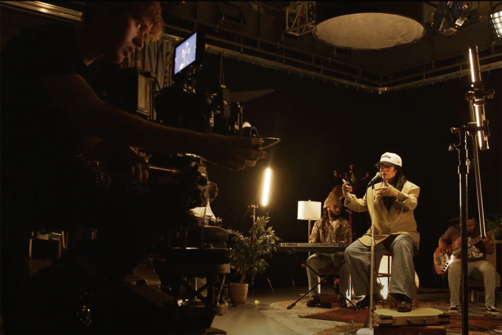 A cameraman sits on a dolly operating a camera while musician Lisa Bello and her band perform live on a set|||